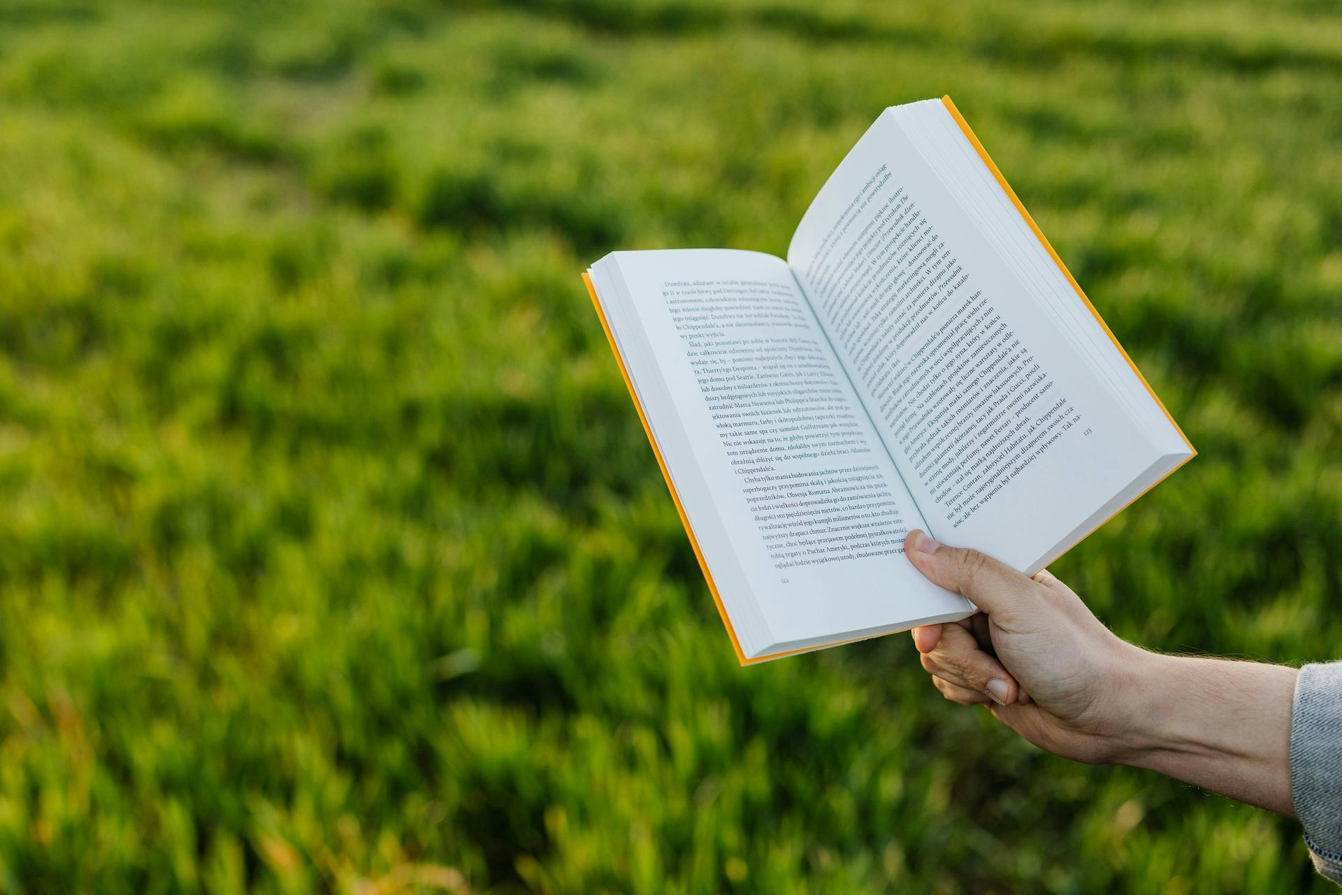 Crop faceless person reading book while sitting on beautiful green lawn in summer countryside at sundown