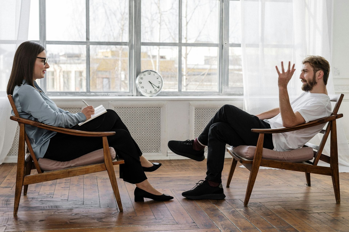 Person in Black Pants and Black Shoes Sitting on Brown Wooden Chair