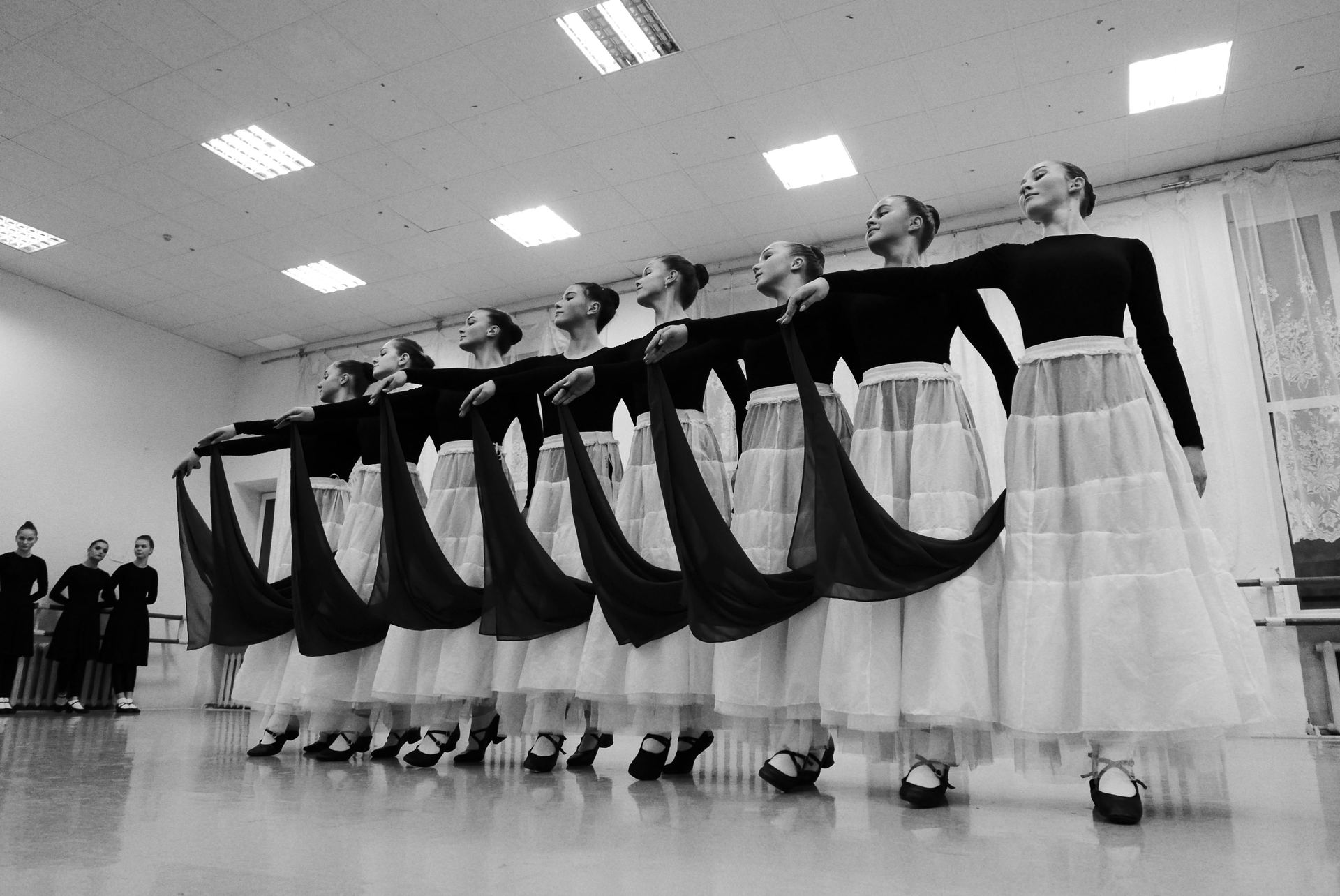 Black and white of female dancers in same wear performing folkloric dance while looking away on floor in light room
