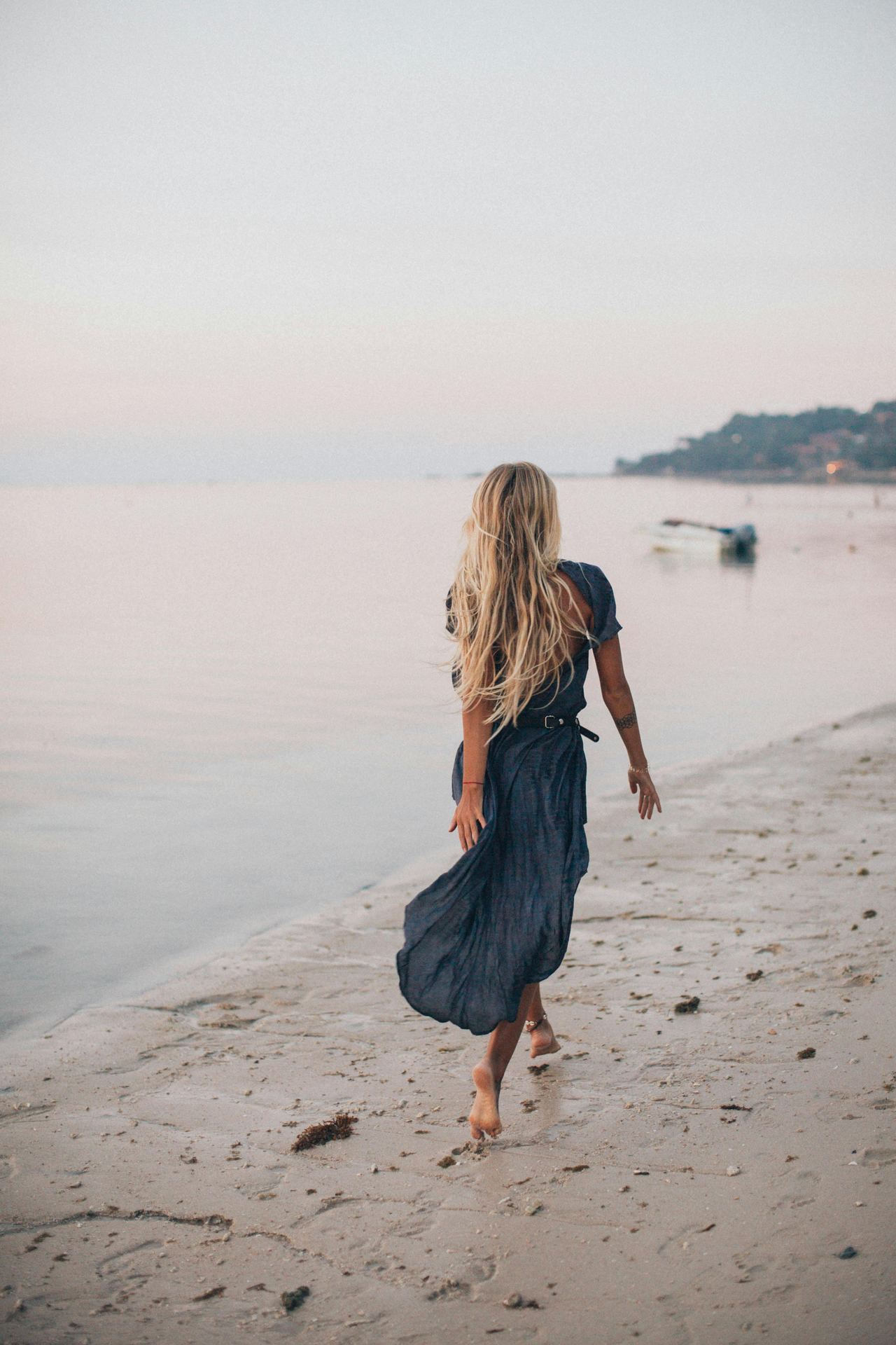 A woman walks barefoot on the seashore at sunset, wearing a blue dress for a serene vacation moment.