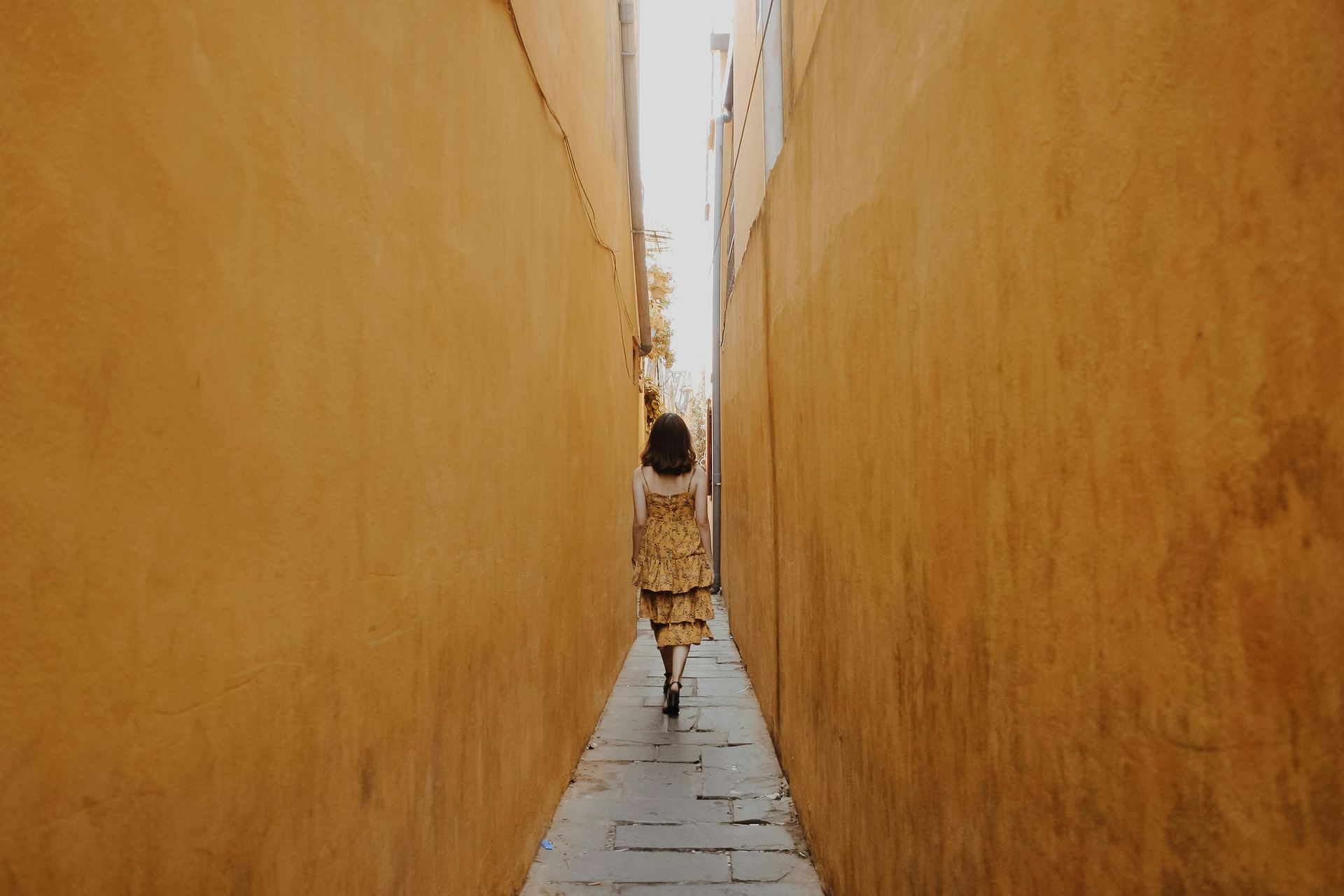 A woman in a yellow dress walks through a narrow alleyway with yellow walls in Há»i An, Vietnam.