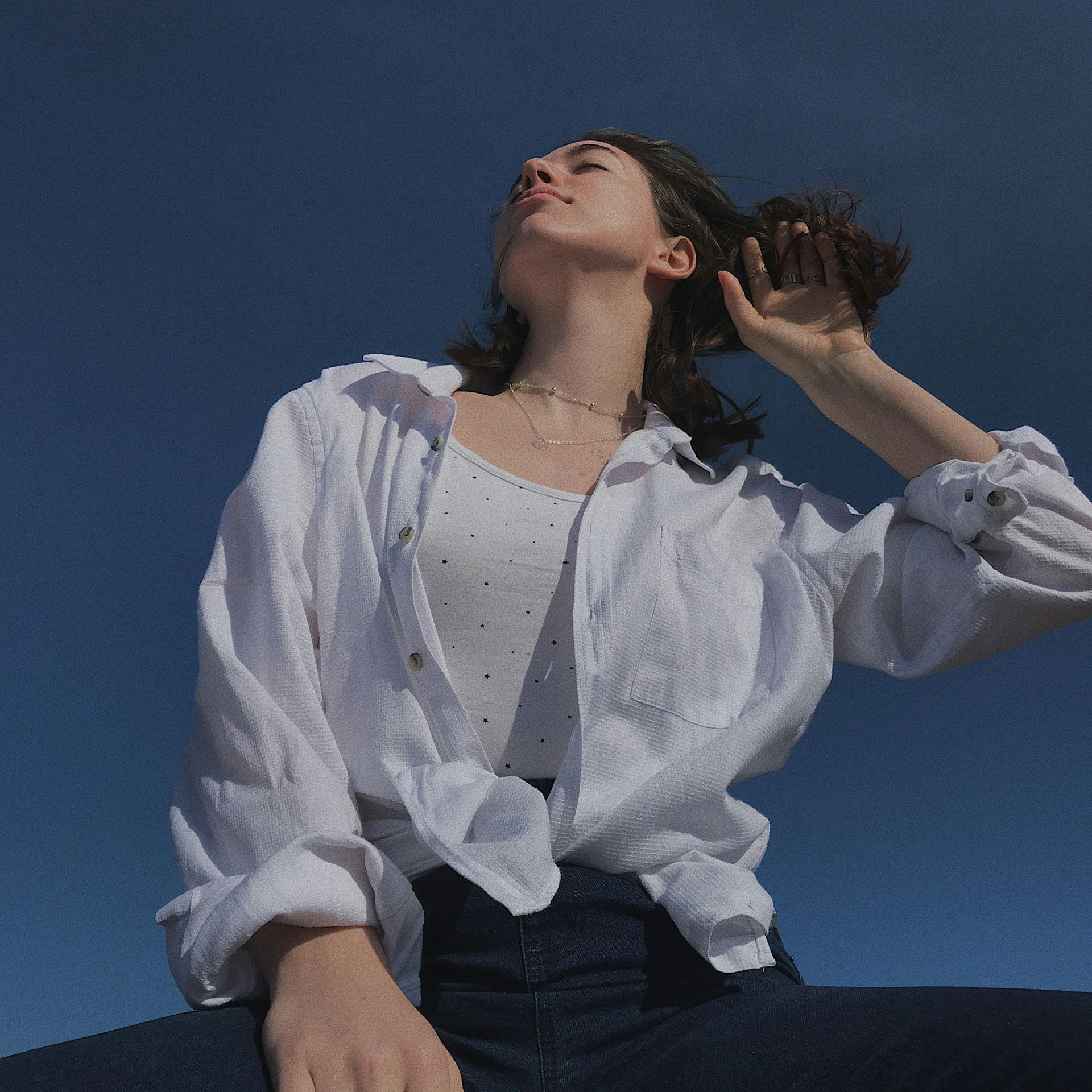 Low angle of young female in casual wear enjoying windy weather and sunlight against blue sky