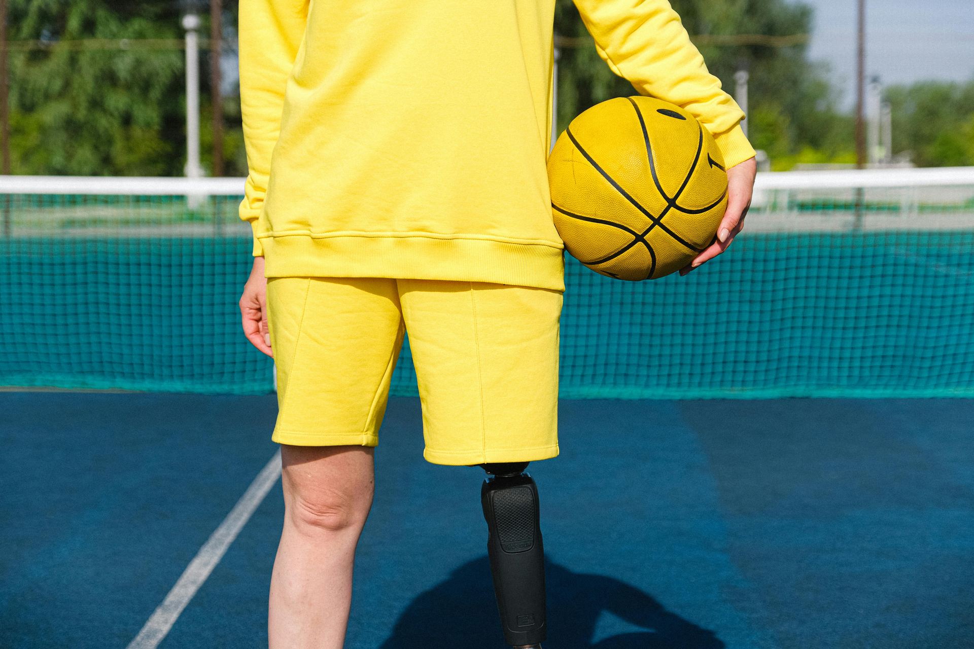 A person with a prosthetic leg stands on a basketball court holding a yellow basketball.