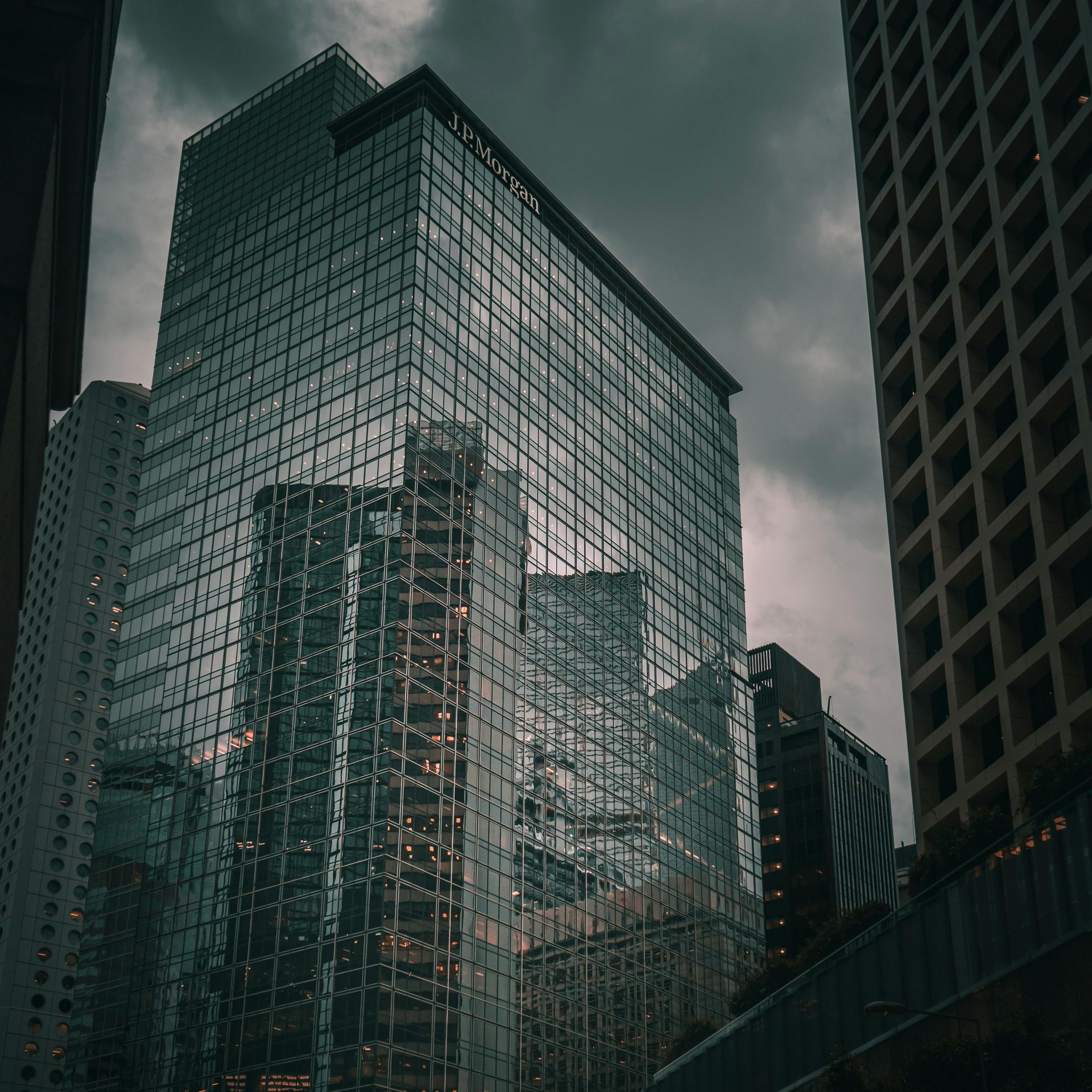From below of tall commercial skyscraper with glass mirrored walls reflecting modern buildings of downtown against gray cloudy sky
