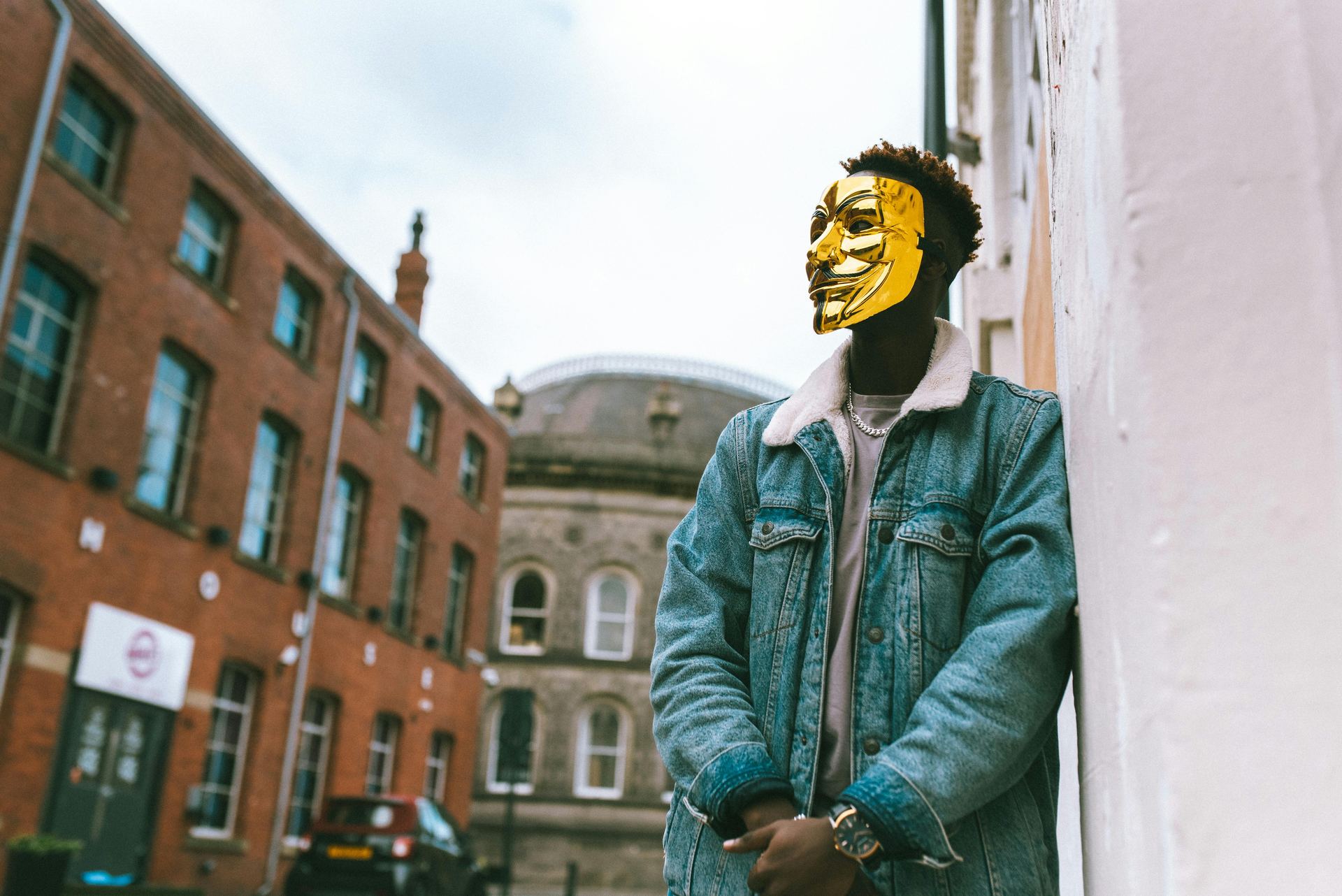 From below of confident black activist in stylish outfit and famous Anonymous mask leaning on building placed in city district in daytime